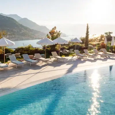 Outdoor swimming pool with chairs and umbrellas, overlooking a body of water and mountains in the background.