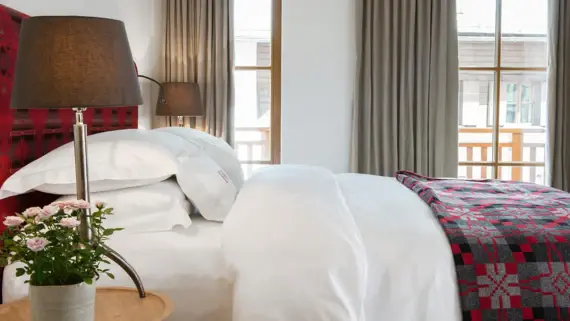 A red upholstered bed with white bed linen and a red bedspread. A bedside table with a lamp and a flower can be seen in the foreground and a floor-to-ceiling window with access to a balcony in the background. 