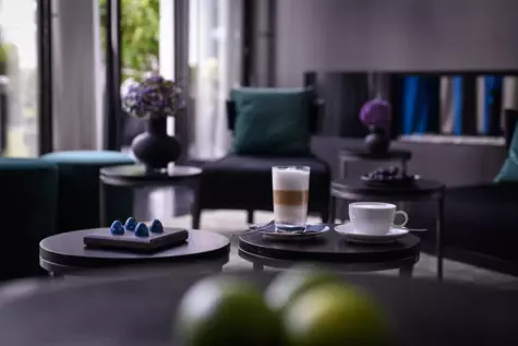 A cream and a latte macchiato are placed on a coffee table in a lounge area elegantly furnished in dark colours. 