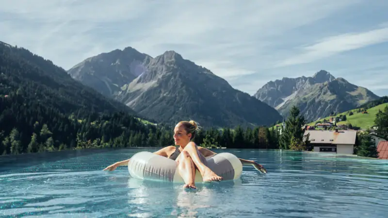 A woman floats on a floating tyre in a pool with mountains in the background.