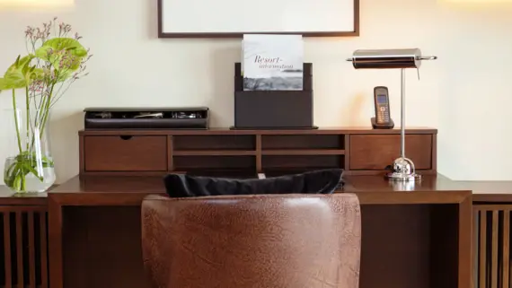 A brown leather armchair stands in front of a dark wood desk. On the desk is a lamp, a telephone, a vase of flowers and several flyers.