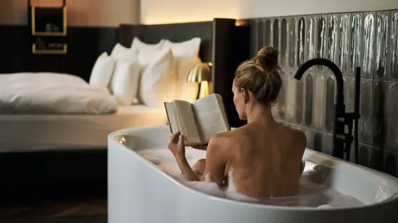 Woman reading a book in a bathtub, in the background a hotel bed with white bed linen.