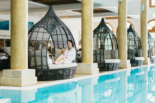 Four lounge seats are placed between columns alongside a pool. A woman in a robe is sitting in one of them enjoying an orange drink. 