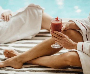 A woman in a robe sitting on a towel is holding a red drink in her hands. 