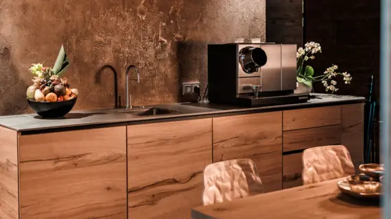 A wooden-look kitchenette with a sink, a coffee machine and a fruit basket. A wooden table with two chairs can be seen in the foreground.