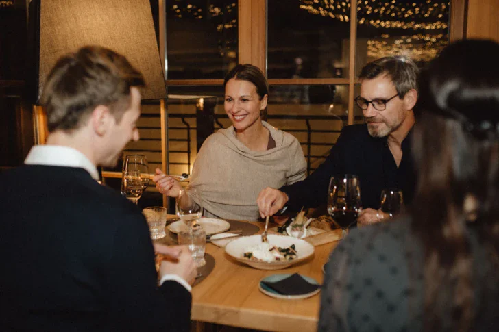 A group of four people sitting around a dining table in a cozy looking restaurant sharing a dessert and drinking wine. 