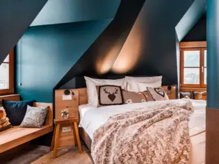 A cozy bed with a brown blanket and three decorative cushions in Alpine style. To the left of the bed is a small wooden bedside table and a seating area with cushions.