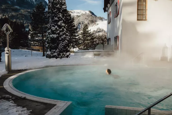 A woman is swimming in a warm steaming outdoor pool of a hotel surrounded by snowy landscapes. 