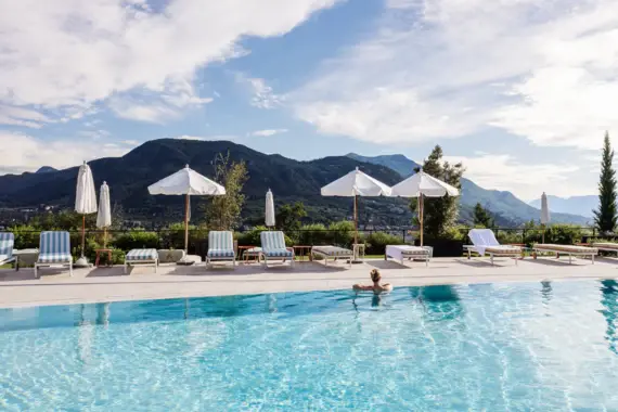 Person leaning on the edge of a pool with parasols, striped deckchairs and mountains in the background.