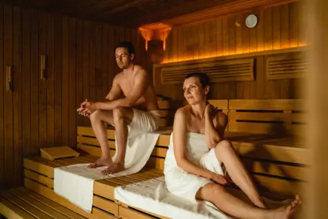 A man and a woman sitting in a wooden sauna with warm orange light. 