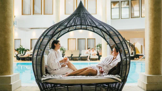 Two woman in robes and with a drink in their hands are sitting in a lounge chair by a pool.