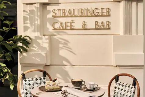 A laid table with two cups of coffee, a piece of cake and a newspaper.