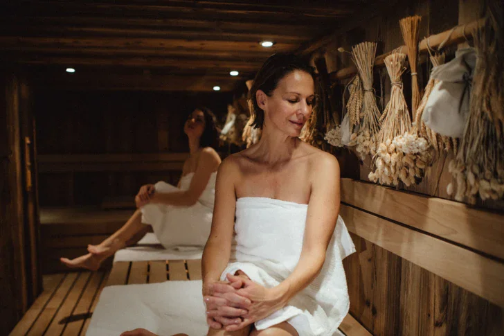 Two woman wrapped in withe towels are sitting in a wooden sauna with bundles of dry herbs and flowers hanging from the wall. 