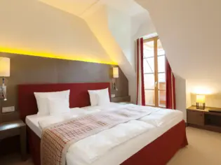 A bedroom with a red upholstered bed on which a narrow plaid bedspread lies. A floor-to-ceiling window can be seen in the background.