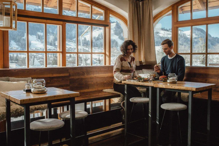 A man and woman sitting at a table in a rustic, cozy looking restaurant with windows in the back, overlooking snowy mountains. 