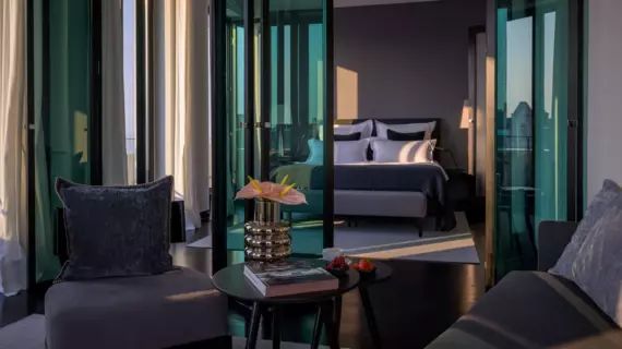 In the foreground are a comfortably upholstered couch and seating options. Flowers are arranged in an abstract silver vase on the small coffee table. A dark green pane of glass separates the front part of the hotel room from the sleeping area, which has a large gray and white bed. 