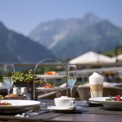 Tisch im Freien mit Geschirr, darunter Teller mit Essen und Getränken mit Bergen im Hintergrund.