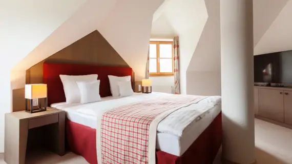 A red upholstered bed stands between a wall and a pillar and there is a plaid bedspread at the foot of the bed. There is a lamp to the left and right of the bed on bedside cabinets. A small window can be seen in the background.