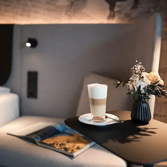 Ein Beistelltisch mit einem Latte Macchiato, einer aufgeschlagenen Zeitschrift und einer Vase mit Blumen neben einem Bett mit gepolstertem Kopfteil vor einer kunstvoll gestalteten Tapete..
