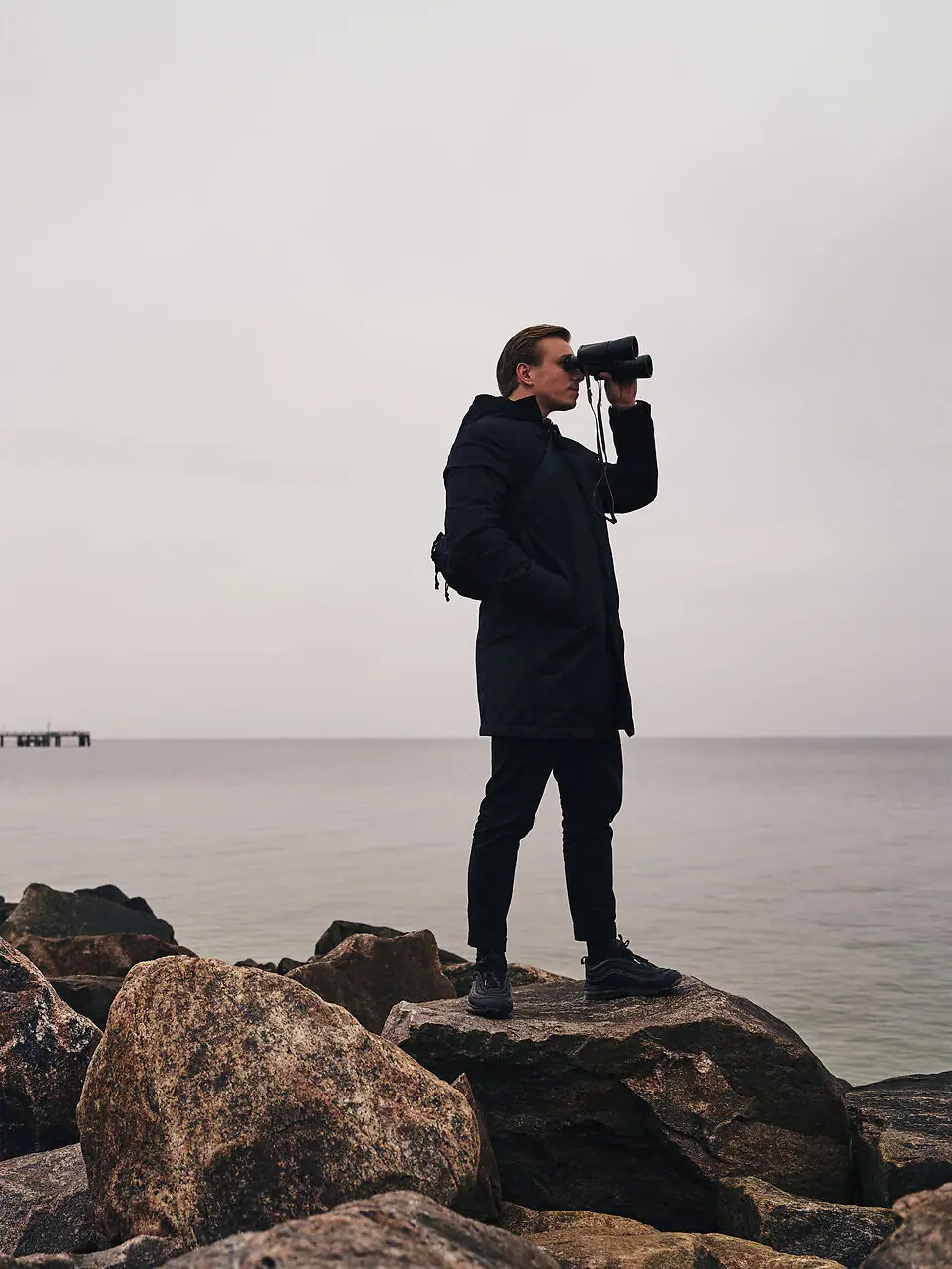 A man is looking through a pair of binoculars at some rocks by the water.