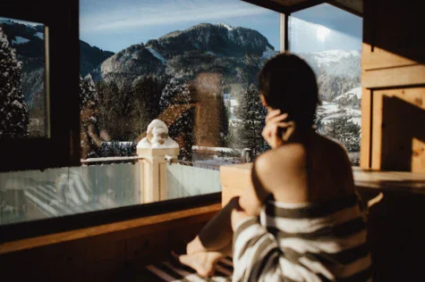 A woman in a sauna overlooking the snowy mountains in the background through a big window front. 