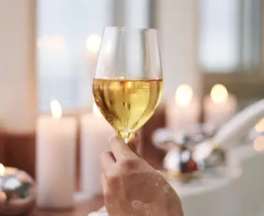  The picture shows a hand holding a wine glass filled with white wine on the edge of a bathtub. The scene radiates a warm and relaxing atmosphere, which is enhanced by the soft candlelight in the background. The blurred contours in the background suggest a luxurious bath. Small drops of water and foam on the hand indicate that the person is enjoying a bath.