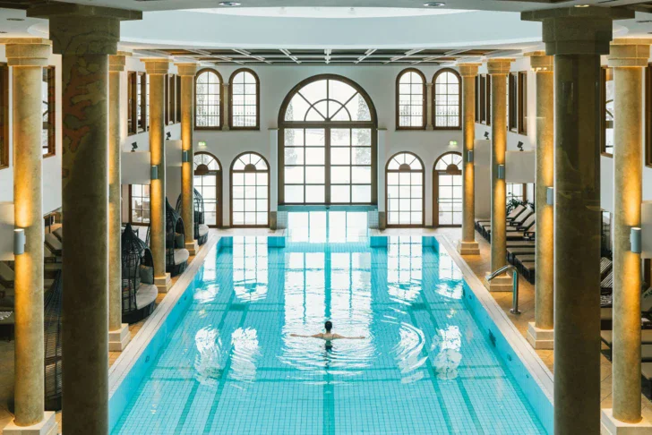 A woman swimming in an indoor pool with illuminated columns and surrounding loungers.