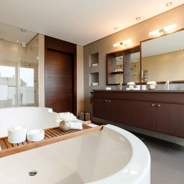 An elegant bathroom with a large, white bathtub and a wood-panelled double washbasin with a mirror hanging above it. Next to the wooden door of the bathroom is the glass shower