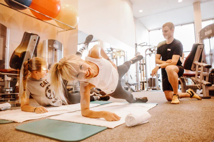 Two women do sports exercises on yoga mats in a fitness room and a trainer watches them in the background.