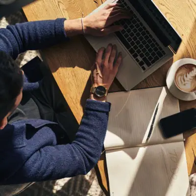 Person, die einen Laptop auf einem Tisch benutzt, daneben steht eine Tasse Cappuccino.