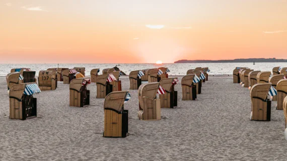 Beach chairs with blue and red and white sunshades are dotted along a quiet beach. The sea is calm and the setting sun bathes the scene in a warm light. 
