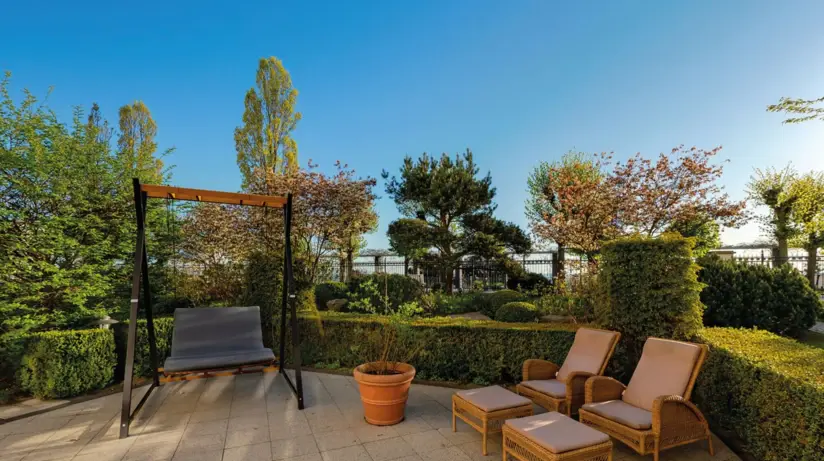 In the outdoor area of the hotel, there are two loungers and a swing in a tiled area. The area behind it is planted with greenery. 