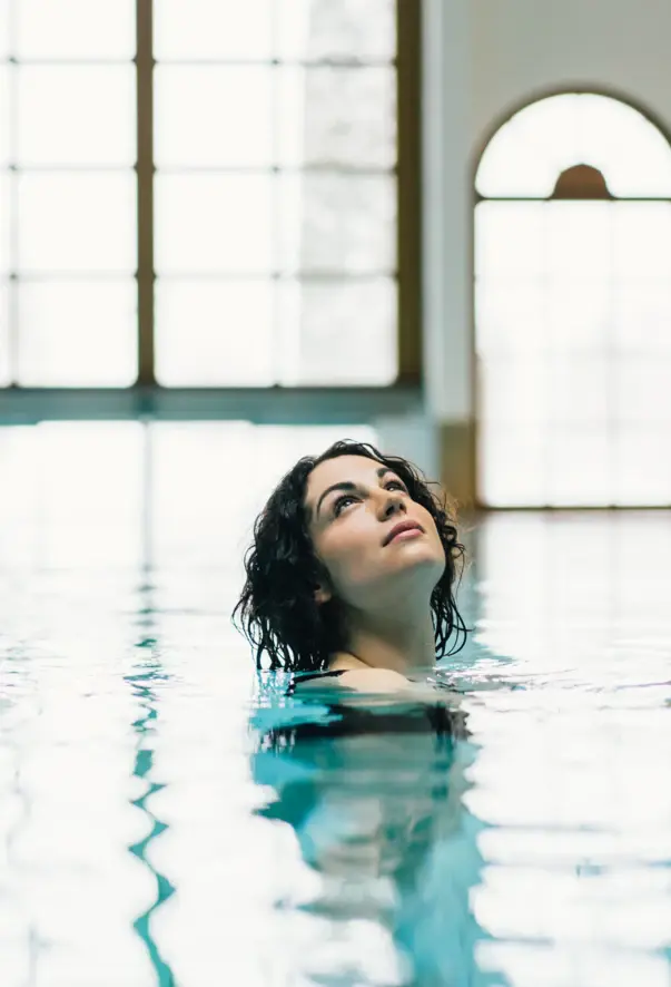 A woman is swinging shoulder deep in a pool looking up to the ceiling. 