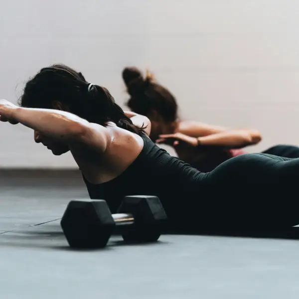 Zwei Frauen liegen mit dem Bauch auf dem Boden und heben ihre Oberkörper an. Im Vordergrund liegt eine Hantel auf dem Boden.