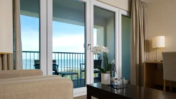 The window front of a hotel room was photographed from a light-colored armchair. In the foreground you can see a dark coffee table with a bottle of water, two glasses and an orchid on it. A balcony with two chairs and a side table can be seen in the background.