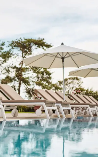 Pool loungers lined up by an outdoor swimming pool under white parasols with trees in the background.