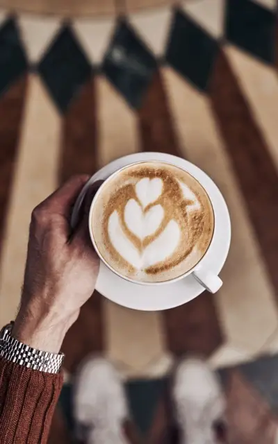 Person holding a cup of cappuccino over a patterned tiled floor.