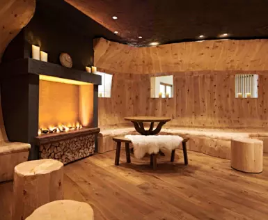 A lounge with fully wooden interior and a fireplace, lighting up the room in warm colors. 