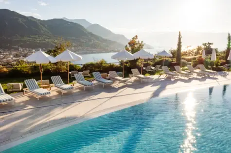 A photorealistic depiction of a luxurious outdoor pool in a hotel complex. Several people relax on loungers and designer seating, some under white umbrellas. Lush greenery, tall cypress trees and flowering shrubs surround the pool area, while a blue sky with light clouds adorns the background. A modern building with terraces and green roofs completes the idyllic setting.