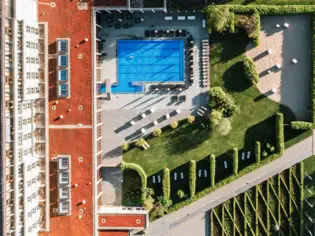 Bird's eye view of the outside of a pool in front of a building with surrounding trees and a road.