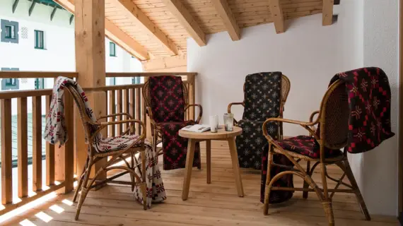 There are four chairs on a balcony made of light-colored wood, each with a blanket over the backrest. In the middle of the chairs is a small wooden table with a book and two drinks.