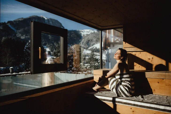 A woman is sitting in wooden sauna with a big glas fron overlooking the snowy mountains and trees. 