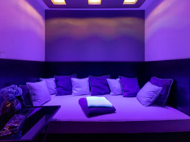 A purple-lit room filled with a large lounge bed full of cushions.