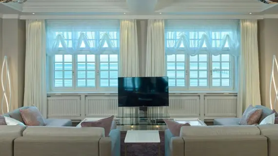 A bright living room in a suite with a window view of the Baltic Sea. The sofas are in neutral tones and feature decorative cushions in shades of pink and purple. Elegant white curtains and transparent, light-colored drapes hang in front of the classic white window frames. Between the sofas is a modern flat-screen TV on a glass stand, surrounded by creative floor lamps that provide cozy lighting.