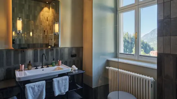 A bathroom with a large window on the right and a washbasin with a mirror hanging over it on the left.