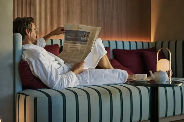 A man in a robe reading a newspaper on a couch indoors.