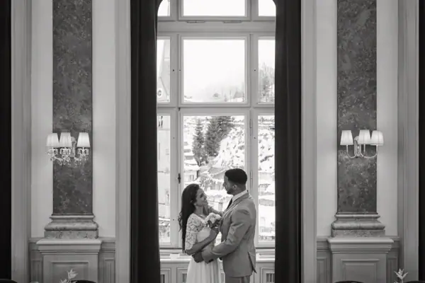 A man and woman standing in a room with a window, wearing formal clothing.
