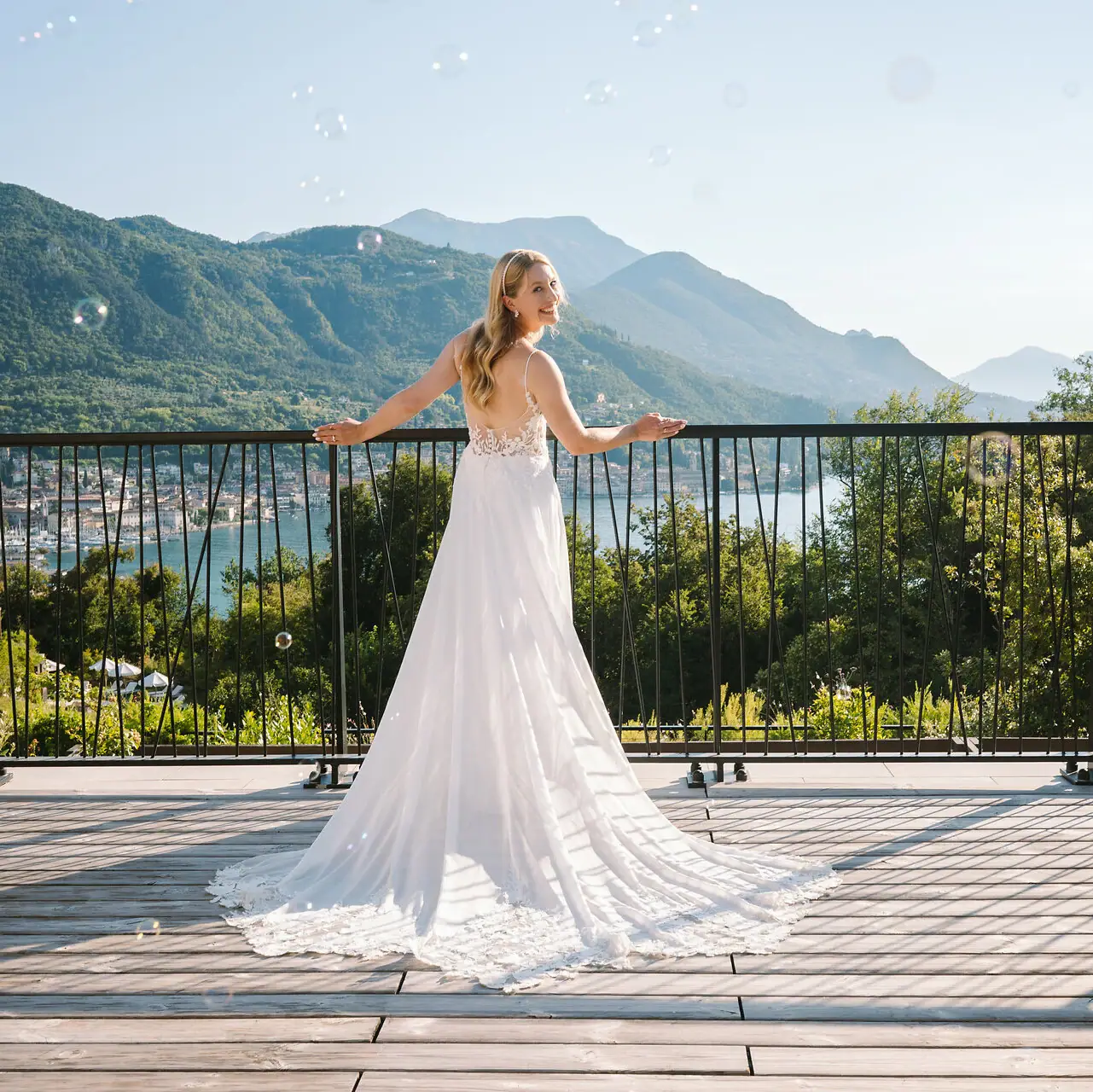 Eine Frau in einem weißen Brautkleid steht auf einem Deck mit einem Geländer, im Hintergrund sind Berge zu sehen.