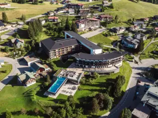 Large building surrounded by grass and trees in a mountain village, surrounded by other alpine-style houses, taken from a bird's eye view.