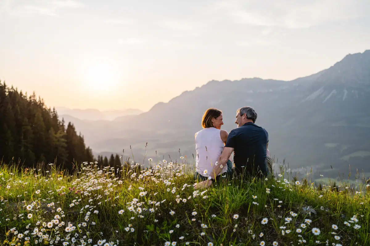 Ein Mann und eine Frau sitzen auf einem Hügel mit Blumen, im Hintergrund sind Berge zu sehen.